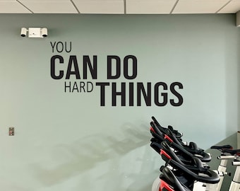 May include: A motivational quote on a wall in a gym, "You can do hard things."  The quote is in black lettering on a light green wall.  There are several stationary bikes in the background.