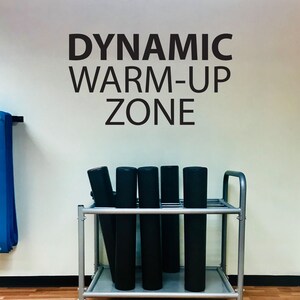 May include: A silver metal cart with black exercise mats stacked on it. The cart is in front of a white wall with the words "DYNAMIC WARM-UP ZONE" written in black.