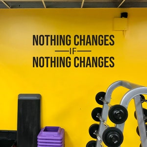 May include: A yellow gym wall with the text "NOTHING CHANGES IF NOTHING CHANGES" in black. A step platform, a stack of purple step risers, and a weight rack with barbells are in the foreground.