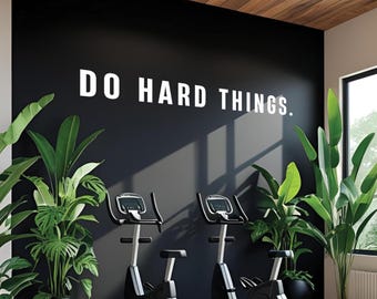 Puede incluir: Un gimnasio moderno con dos bicicletas estáticas. La pared negra muestra el texto blanco "DO HARD THINGS". Plantas verdes en macetas están colocadas en la habitación. El techo es de madera.