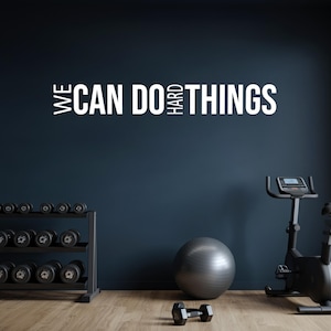 May include: A home gym with a dark blue wall featuring the white text "WE CAN DO HARD THINGS." The room includes a dumbbell rack, exercise bike, exercise ball, and a single dumbbell on the floor. The floor is made of wood.