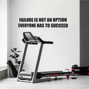 Könnte beinhalten: Ein schwarz-weißes Motivationszitat an einer weißen Wand mit der Aufschrift "Scheitern ist keine Option, jeder muss erfolgreich sein." Im Vordergrund steht ein schwarzes und silbernes Laufband.