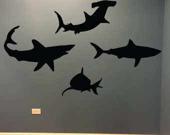 May include: A wall with five black silhouette decals of sharks and hammerhead sharks. The sharks are facing different directions and are all different sizes.