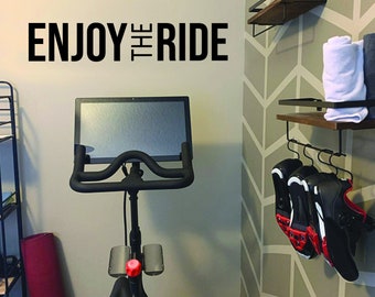 May include: A black stationary exercise bike with a screen displaying a workout. The bike is in a room with a gray wall with a geometric pattern and a shelf with shoes and towels. The wall also has a black vinyl decal that reads "ENJOY THE RIDE".