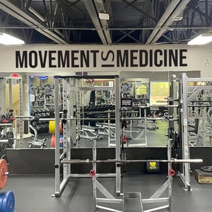 Peut inclure: Une salle de sport avec une pancarte indiquant "MOVEMENT IS MEDICINE" au-dessus d'un miroir refl&eacute;tant une zone de musculation avec un rack &agrave; squat et des poids.