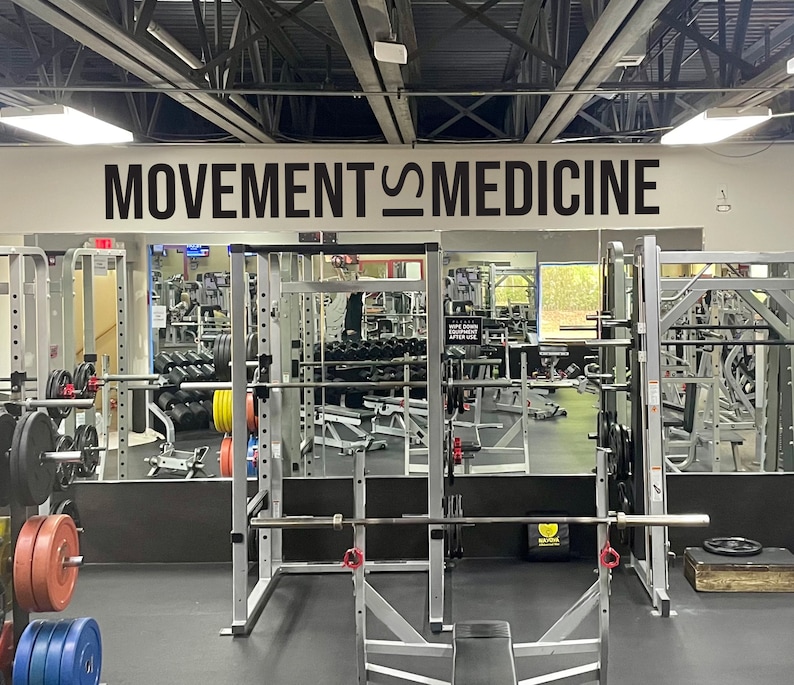 Peut inclure: Une salle de sport avec une pancarte indiquant "MOVEMENT IS MEDICINE" au-dessus d'un miroir refl&eacute;tant une zone de musculation avec un rack &agrave; squat et des poids.