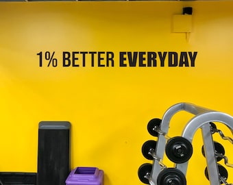 Peut inclure: Un mur jaune avec le texte "1% BETTER EVERYDAY" en lettres noires. Il y a un rack de musculation avec plusieurs barres dans le coin inférieur droit de l'image.
