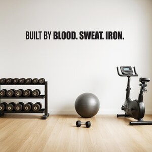 May include: A home gym setup with the text "BUILT BY BLOOD. SWEAT. IRON." on the wall. The room includes a dumbbell rack with multiple dumbbells, a stationary exercise bike, a gray exercise ball, and a single dumbbell on the floor.