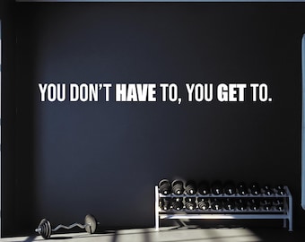 Peut inclure: Un mur noir avec le texte blanc "YOU DON'T HAVE TO, YOU GET TO." Un support en métal blanc contient plusieurs haltères noirs, et une barre de musculation noire avec une barre ondulée repose au sol au premier plan.