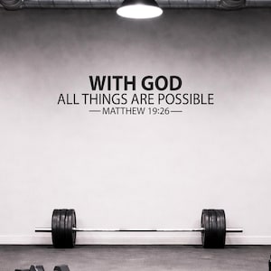 Può includere: Un adesivo murale nero con il testo WITH GOD ALL THINGS ARE POSSIBLE — MATTHEW 19:26 — in un carattere sans-serif pulito. La scritta è applicata su una superficie murale liscia e chiara, posizionata sopra un bilanciere in metallo con dischi neri.
