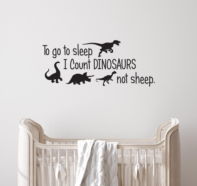 May include: A white crib with a white blanket in it. The wall behind the crib has a black vinyl decal that says "To go to sleep I count dinosaurs not sheep." There are four dinosaur silhouettes in the decal.