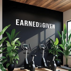 Peut inclure: Une salle de sport moderne avec deux vélos d'exercice stationnaires. Les vélos sont noirs et argentés, placés devant un mur noir avec le texte blanc "EARNED NOT GIVEN". Des plantes en pot ajoutent de la verdure. Le plafond est en bois.