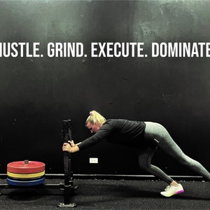 May include: A woman in black and grey workout clothes is pushing a weight sled. The sled has a stack of red, yellow, and blue weight plates. The text "HUSTLE. GRIND. EXECUTE. DOMINATE." is written on the wall behind her.