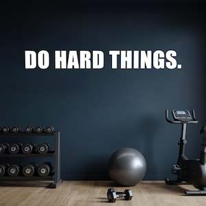 May include: A motivational fitness studio scene with the text "DO HARD THINGS" in white against a dark blue wall. The room features a dumbbell rack, exercise bike, exercise ball, and a single dumbbell on a wooden floor.