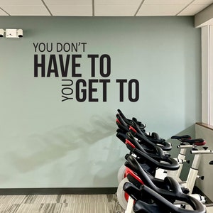 May include: A row of stationary exercise bikes with black seats and handlebars, and red accents. The bikes are in a gym setting, with a light green wall featuring the text "YOU DON'T HAVE TO YOU GET TO".