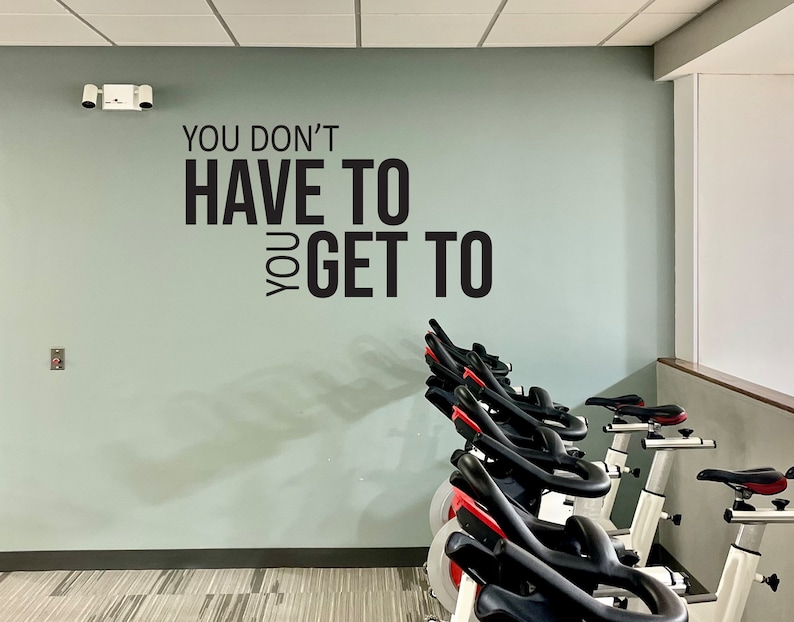 May include: A row of stationary exercise bikes with black seats and handlebars, and red accents. The bikes are in a gym setting, with a light green wall featuring the text "YOU DON'T HAVE TO YOU GET TO".