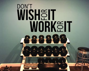 Peut inclure: Une citation de motivation sur un mur bleu qui dit "Ne le souhaite pas, travaille pour ça". Un rack à poids avec des haltères noirs est au premier plan.