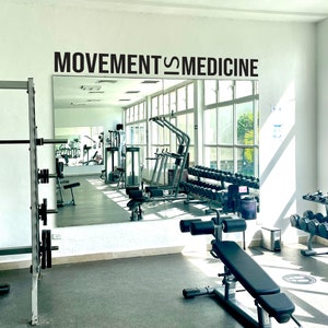 Peut inclure: Une salle de sport avec un grand miroir sur le mur qui dit "MOVEMENT IS MEDICINE". Il y a des bancs de musculation, des halt&egrave;res et d'autres &eacute;quipements de sport dans la salle de sport.