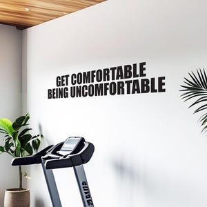 May include: A treadmill with a digital display is positioned in front of a white wall. The wall features the text "GET COMFORTABLE BEING UNCOMFORTABLE" in bold black letters. A potted plant is on the left, and a palm frond is on the right.
