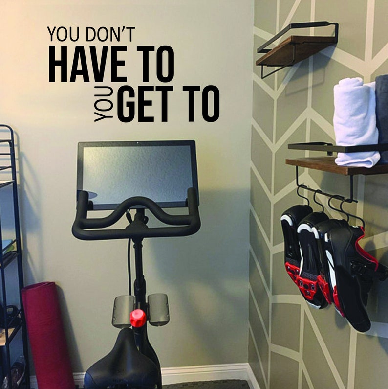 May include: An exercise bike with a large screen is positioned in front of a wall with the text "YOU DON'T HAVE TO YOU GET TO". A red yoga mat leans against the wall. Shelves hold towels and cycling shoes.