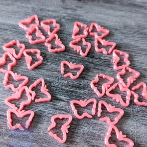 May include: A collection of pink plastic butterfly-shaped charms. The charms are arranged in a circle on a wooden surface.