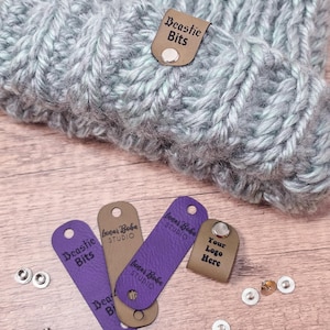 May include: A close-up of a gray and blue knitted beanie with a brown leather tag that says "Beastie Bits". There are also several leather tags with the text "Lunar Boba Studio" and "Your Logo Here".