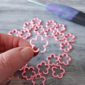 Puede incluir: Marcadores de punto de ganchillo de plástico en forma de flor de color rosa. Hay 15 marcadores en la imagen.