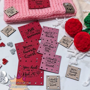 Op de afbeelding: Een roze en oranje gebreide muts met verschillende kleine, vierkante labels. De labels zijn rood, roze en bruin, met zinnen als "Epic Loot Drop" en "You're my Favorite Quest". Sommige labels hebben decoratieve gaatjes.