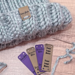 Puede incluir: Primer plano de un gorro de punto gris claro con una etiqueta de cuero marrón que dice "Lunar Boba STUDIO". Varias etiquetas con texto están al lado del sombrero, incluyendo "Beastie Bits" y "Your Logo Here".