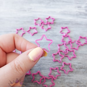 May include: A close-up of a pink star-shaped plastic ring. The ring is being held by a hand with yellow and pink nail polish. There are several other pink star-shaped rings scattered on a light wood surface.