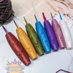 May include: A collection of seven crochet hooks in various colors, including red, gold, green, blue, purple, pink, and white. Each hook has a metallic hook end and a wooden handle. The handles are engraved with "NC" and a number or letter. A ball of yarn is in the background.
