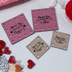 Op de afbeelding: Vier kleine, vierkante labels met de zinnen "You're My Favorite Player 1" en "Player Two Has Entered the Game" erop gedrukt. De labels zijn roze en bruin, met kleine gaatjes in elke hoek. Hartvormige glitterversieringen zijn verspreid.