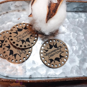 May include: Circular wooden craft pieces with the text "NERDOPRENEUR CREATIONS" in black. Each piece features a black burst design and small holes around the edge. A cotton boll rests on a galvanized metal tray.