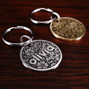 Puede incluir: Dos chapas para perros grabadas en plata y oro con llaveros. La chapa de plata dice "Olivia" y la chapa de oro tiene un dise&ntilde;o floral.