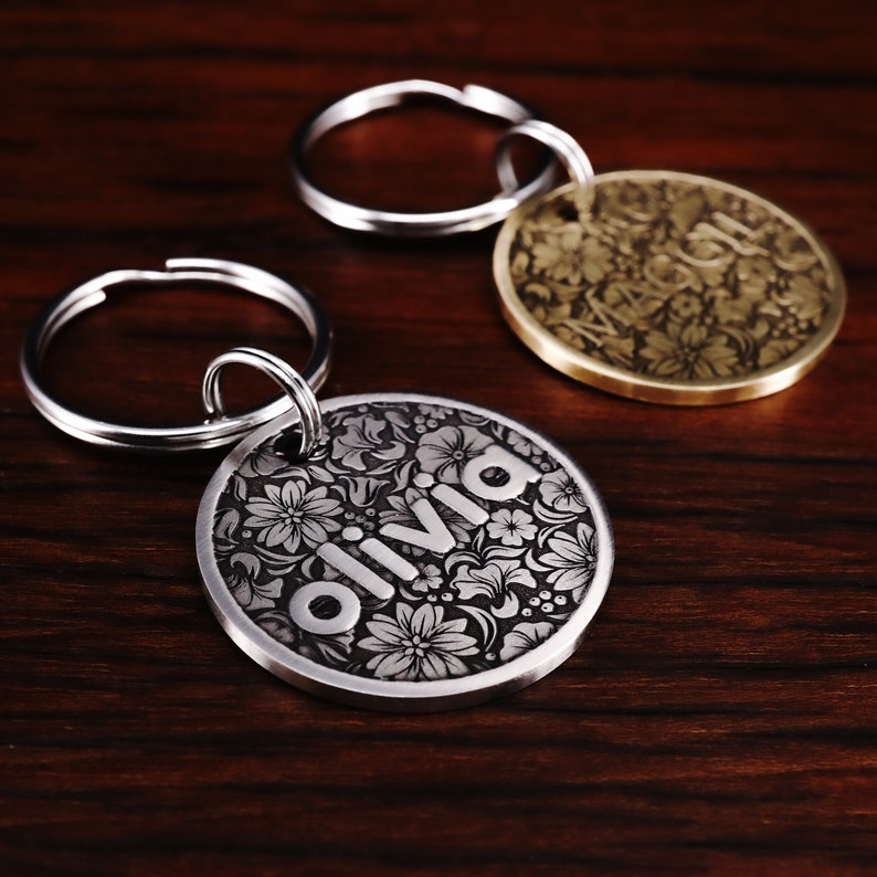 Puede incluir: Dos chapas para perros grabadas en plata y oro con llaveros. La chapa de plata dice "Olivia" y la chapa de oro tiene un dise&ntilde;o floral.
