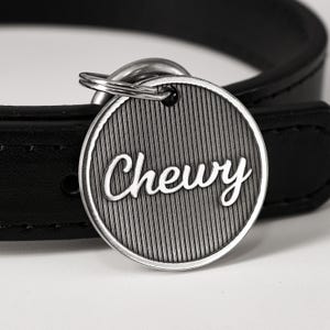 May include: A circular silver-toned metal pet tag featuring a vertical ribbed texture and the name Chewy in a raised script font. The tag is attached to a split ring and rests against a smooth black leather collar with visible stitching.