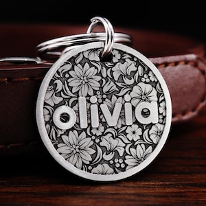 Puede incluir: Chapa de perro de plata con un dise&ntilde;o floral y el nombre "Olivia" grabado en ella.