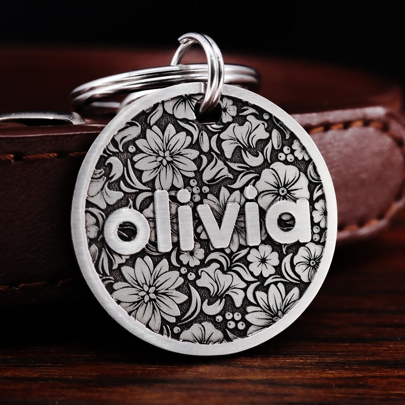 Puede incluir: Chapa de perro de plata con un dise&ntilde;o floral y el nombre "Olivia" grabado en ella.