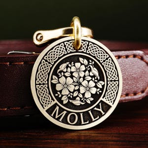 May include: A round, gold-coloured dog tag with a Celtic knot border and a floral design in the centre. The tag is engraved with the name "MOLLY".