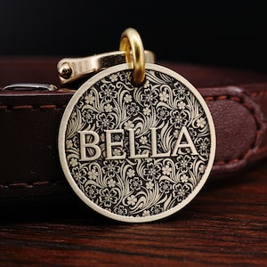 May include: A round brass dog tag with a floral pattern and the name "BELLA" engraved in the center.