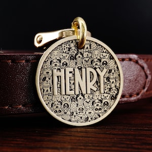 Puede incluir: Chapa de perro dorada con un patr&oacute;n de doodle de perros en blanco y negro y el nombre "HENRY" grabado en ella.