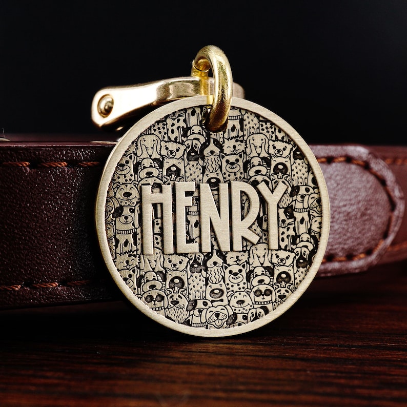Puede incluir: Chapa de perro dorada con un patr&oacute;n de doodle de perros en blanco y negro y el nombre "HENRY" grabado en ella.