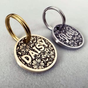 May include: Two engraved dog tags with floral designs. The tag on the left says "DAISY" and is brass. The tag on the right says "ROXY" and is silver.