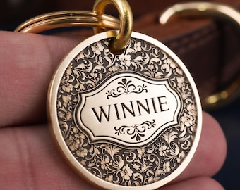 Peut inclure: Une médaille pour animaux de compagnie ronde et dorée avec le nom "WINNIE" gravé dans un cadre décoratif. La médaille présente un motif floral orné sur les bords et comprend un anneau fendu doré pour l'attache.