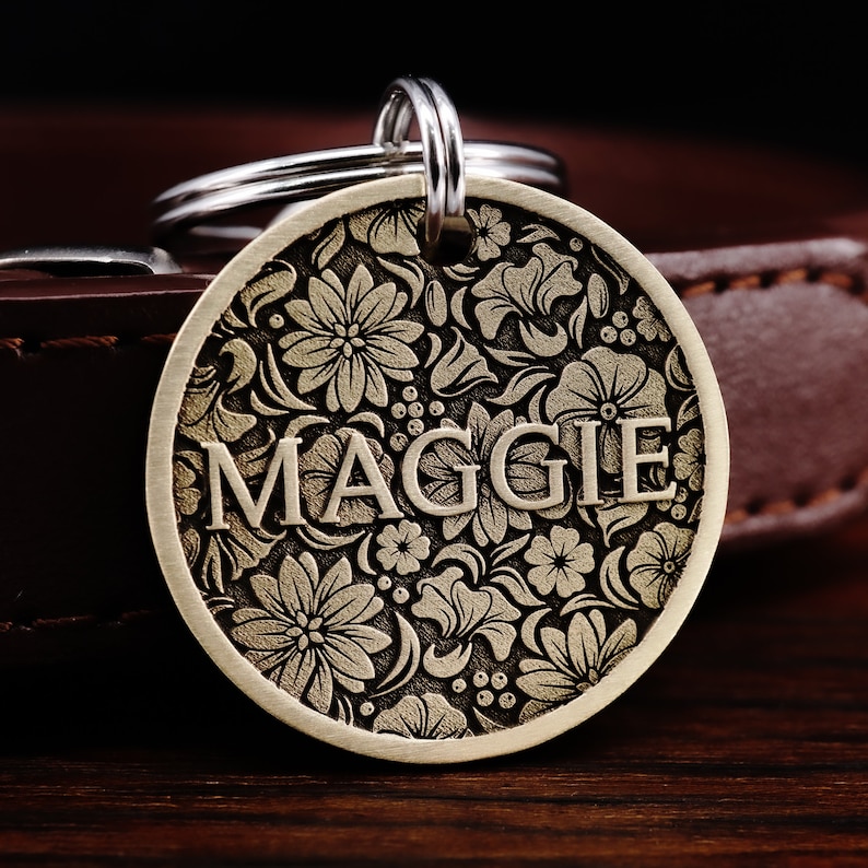 Puede incluir: Una chapa redonda de perro de lat&oacute;n con un dise&ntilde;o floral y el nombre "MAGGIE" grabado en ella. La chapa est&aacute; unida a un llavero de plata.