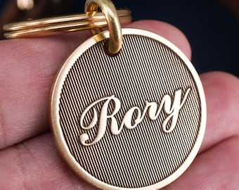 Peut inclure: Une médaille ronde dorée pour animaux de compagnie avec le nom "Rory" en écriture élégante. La médaille présente un motif texturé à lignes diagonales et comprend un porte-clés doré.