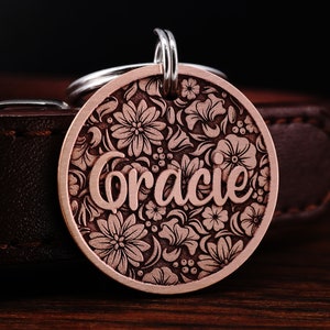 Puede incluir: Una placa de perro de cobre con un dise&ntilde;o floral y el nombre "Gracie" grabado en ella. La placa est&aacute; unida a un collar de cuero.