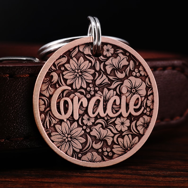 Puede incluir: Una placa de perro de cobre con un dise&ntilde;o floral y el nombre "Gracie" grabado en ella. La placa est&aacute; unida a un collar de cuero.