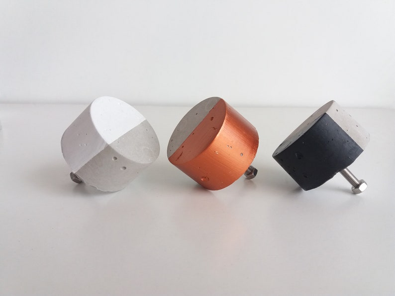 May include: Three concrete cabinet knobs with a split design. The knobs are painted white, copper, and black.