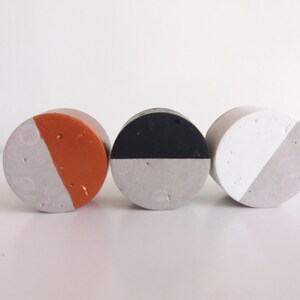 May include: Three round concrete coasters with a half-circle design. The coasters are painted in different colours: orange, black, and white.